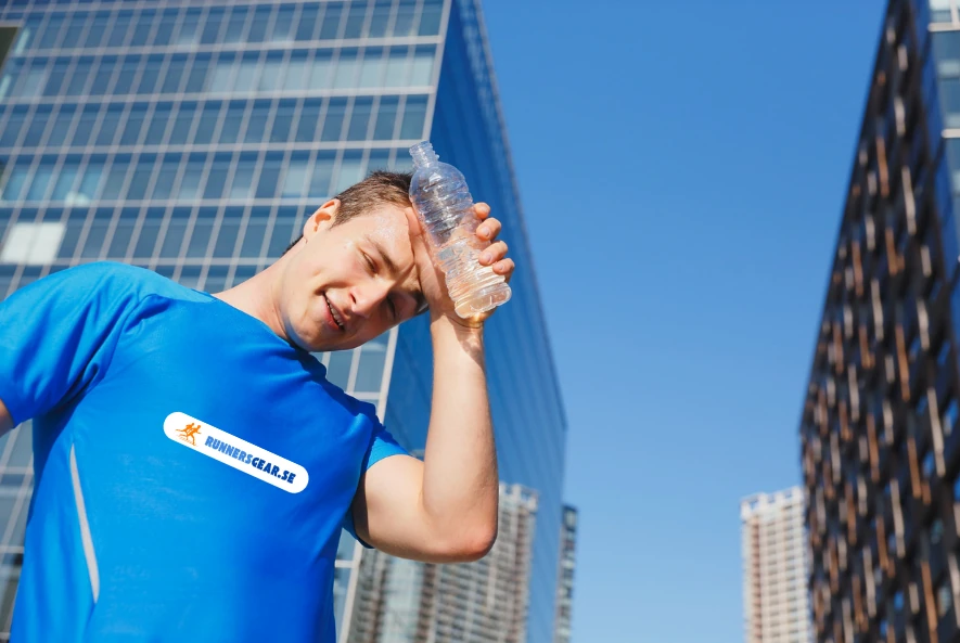 En svettig löpare håller en kall flaska vatten mot pannan, känner sig lite svimfärdig på grund av elektrolytförlust, har en blå t-shirt med trycket RunnersGear.se tryckt mitt på tröjan, i bakgrunden syns en skyskrapa